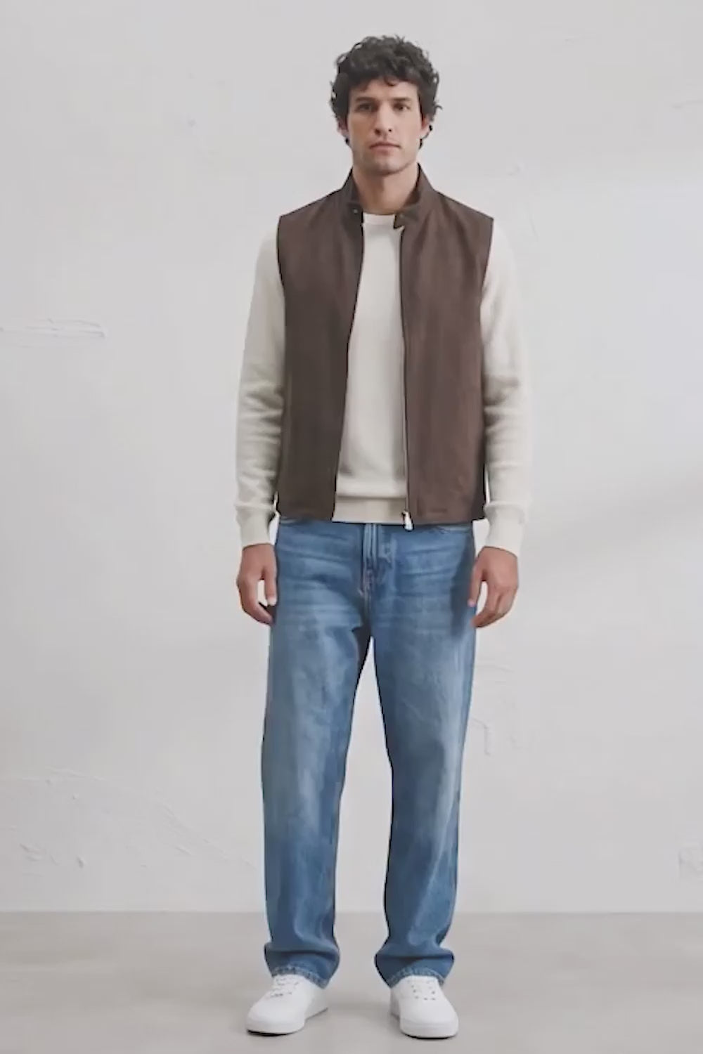 A man modeling a brown sleeveless zippered linen vest over a cream long-sleeve shirt, paired with blue jeans and white sneakers, standing against a white studio wall.