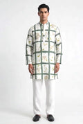 A man modeling a white long-sleeve kurta with a green grid pattern and small yellow floral eco-print accents, paired with white trousers and black shoes, against a light gray studio background.