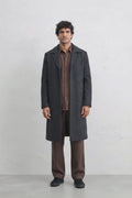 A man modeling a dark charcoal gray linen overcoat over a brown linen shirt and matching brown trousers, standing against a light gray studio background.