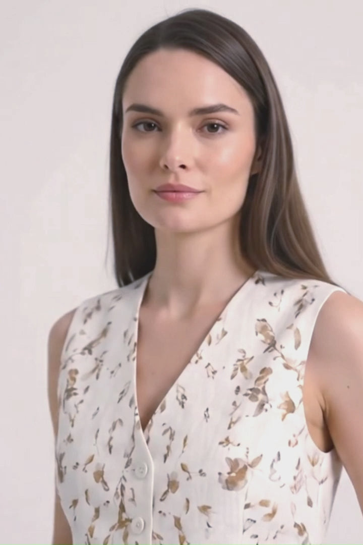 A woman modeling a sleeveless white vest with a gold abstract botanical-print, paired with a white skirt, against a light gray studio background.