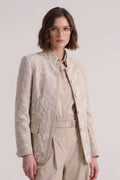 A model stands against a light gray studio background wearing a beige and cream embroidred linen jacket over a matching beige shirt and wide-leg trousers, paired with beige sneakers.