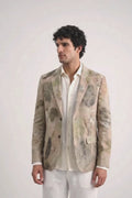 A man modeling a light beige and white abstract-patterned eco-print blazer over a white shirt and white trousers, standing against a plain white studio background.