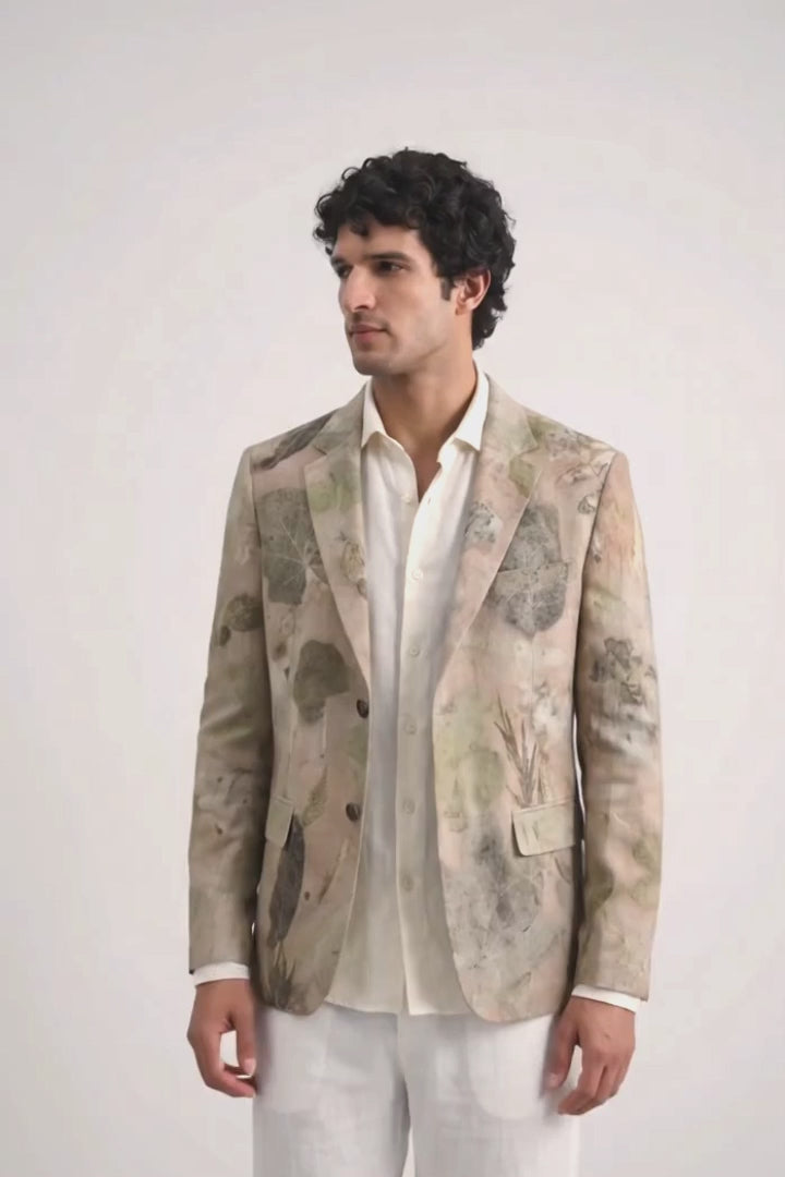 A man modeling a light beige and white abstract-patterned eco-print blazer over a white shirt and white trousers, standing against a plain white studio background.