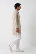 A man modeling a light beige linen kurta with a buttoned mandarin collar, paired with white trousers and white sneakers, standing against a plain white studio background.