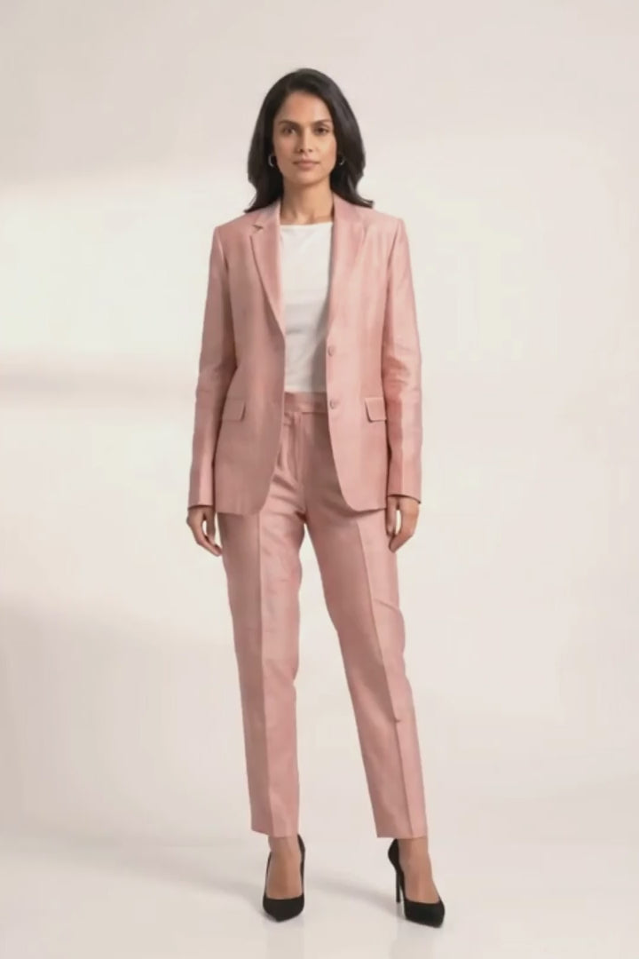 A woman modeling a dusty pink tailored suit set consisting of a silk blazer and matching trousers, styled with a white top and black heels against a light gray studio background.