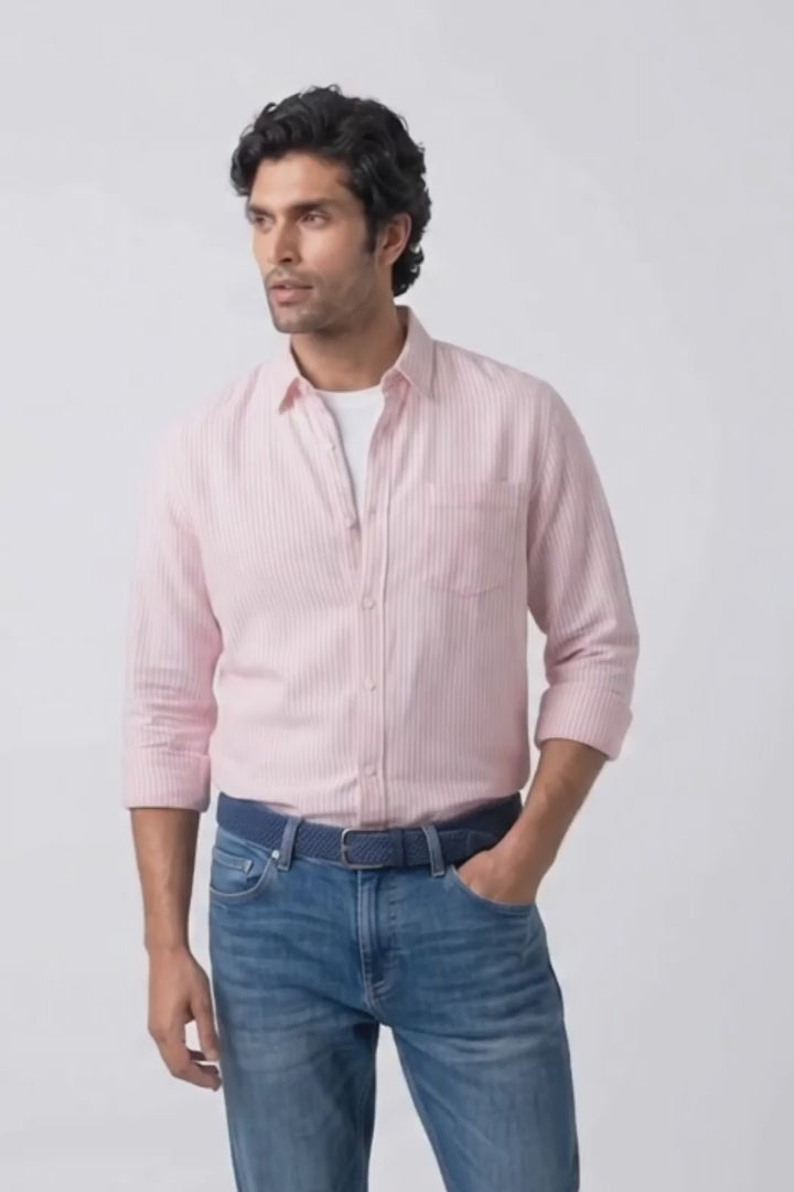 A man modeling a light pink long-sleeve button-down linen shirt and blue jeans against a plain light gray studio background.