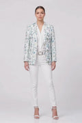 A model stands against a light gray studio background wearing a white button-up shirt, white pants, a white belt, and a light blue and white floral patterned linen blazer, along with white heeled sandals.