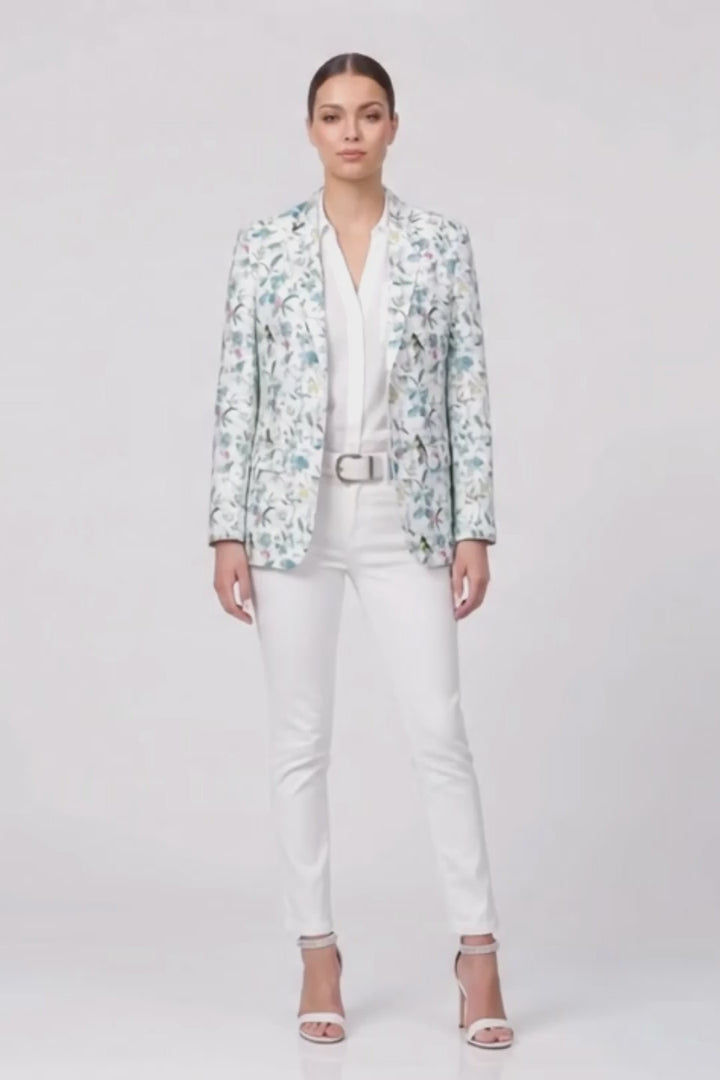 A model stands against a light gray studio background wearing a white button-up shirt, white pants, a white belt, and a light blue and white floral patterned linen blazer, along with white heeled sandals.