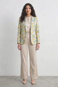 A woman stands against a plain light gray studio wall wearing a beige satin-like blouse and wide-leg beige trousers, with a patterned silk blazer draped over her shoulders. The blazer has a yellow and mint green abstract floral or leaf print.