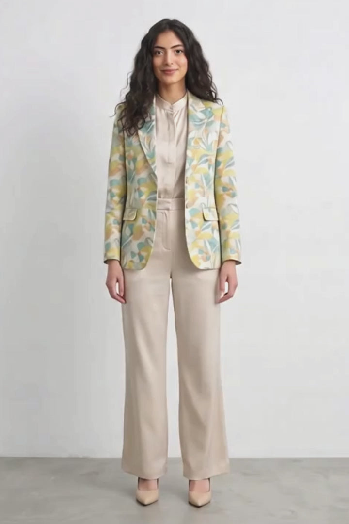 A woman stands against a plain light gray studio wall wearing a beige satin-like blouse and wide-leg beige trousers, with a patterned silk blazer draped over her shoulders. The blazer has a yellow and mint green abstract floral or leaf print.