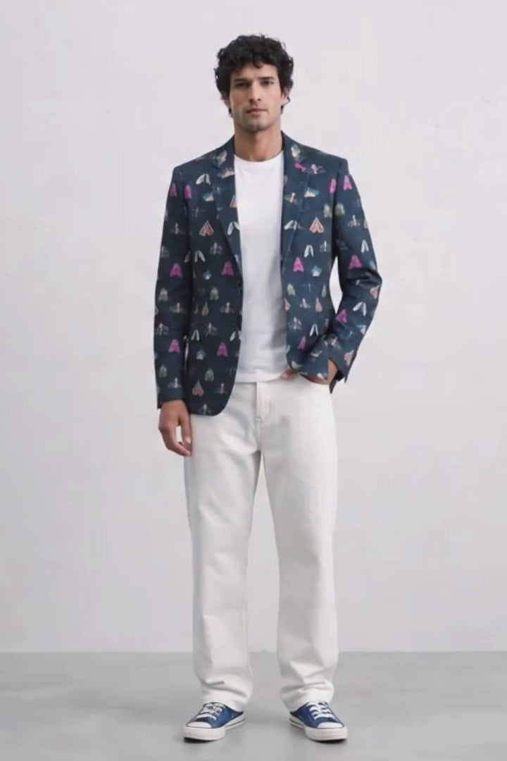 A man modeling a navy blue blazer with a colorful geometric pattern, worn over a white t-shirt and white jeans, paired with blue sneakers, against a plain white studio background.