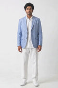 A man modeling a light blue linen-blend blazer over a white shirt and white trousers, standing against a plain white studio background.