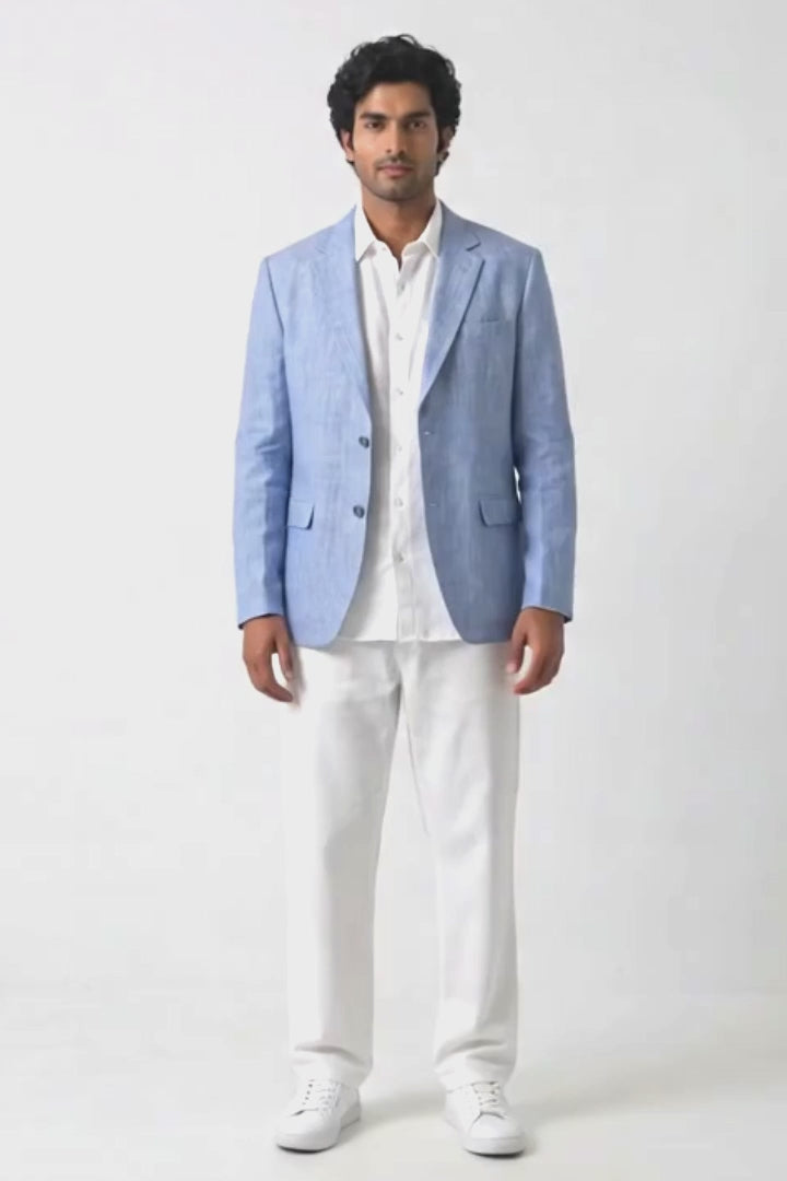 A man modeling a light blue linen-blend blazer over a white shirt and white trousers, standing against a plain white studio background.