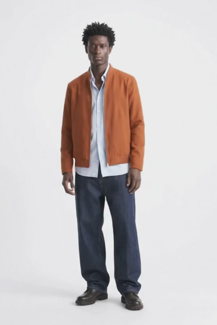 A man modeling a rust-orange linen jacket over a white button-down shirt and loose dark blue jeans, standing against a plain white studio background.