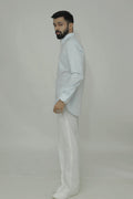 A man standing, wearing a linen shirt with a cut-away collar, long sleeves, and a single-button cuff, paired with light-colored trousers.