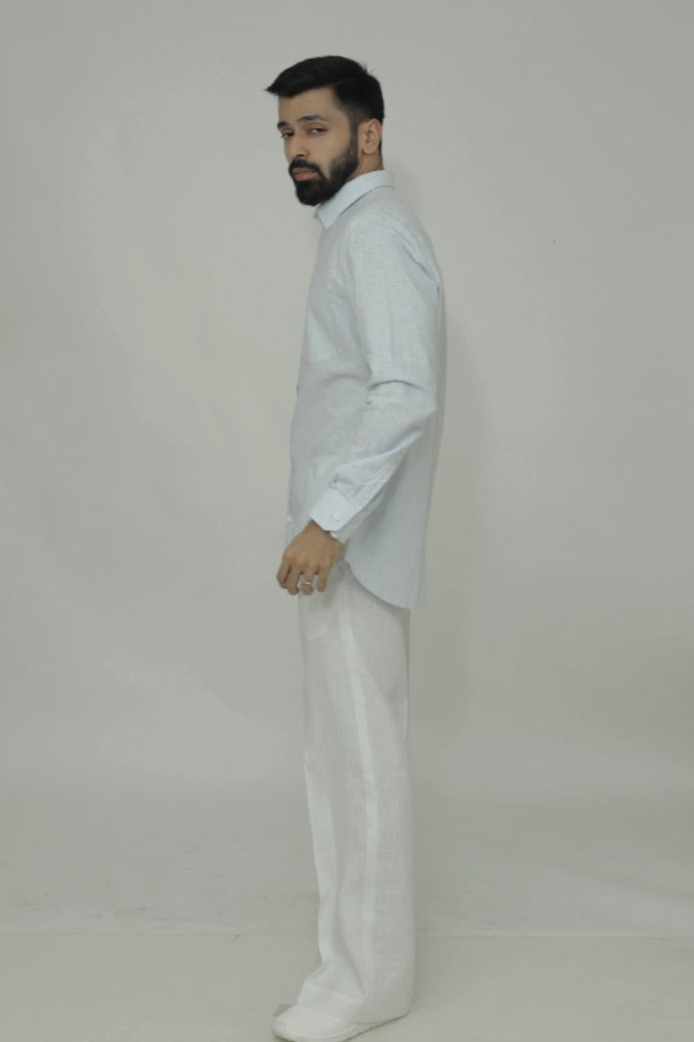 A man standing, wearing a linen shirt with a cut-away collar, long sleeves, and a single-button cuff, paired with light-colored trousers.