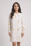 A woman modeling a white long-sleeve shift linen dress with a yellow and orange eco-print, paired with white sneakers, against a light gray studio background.