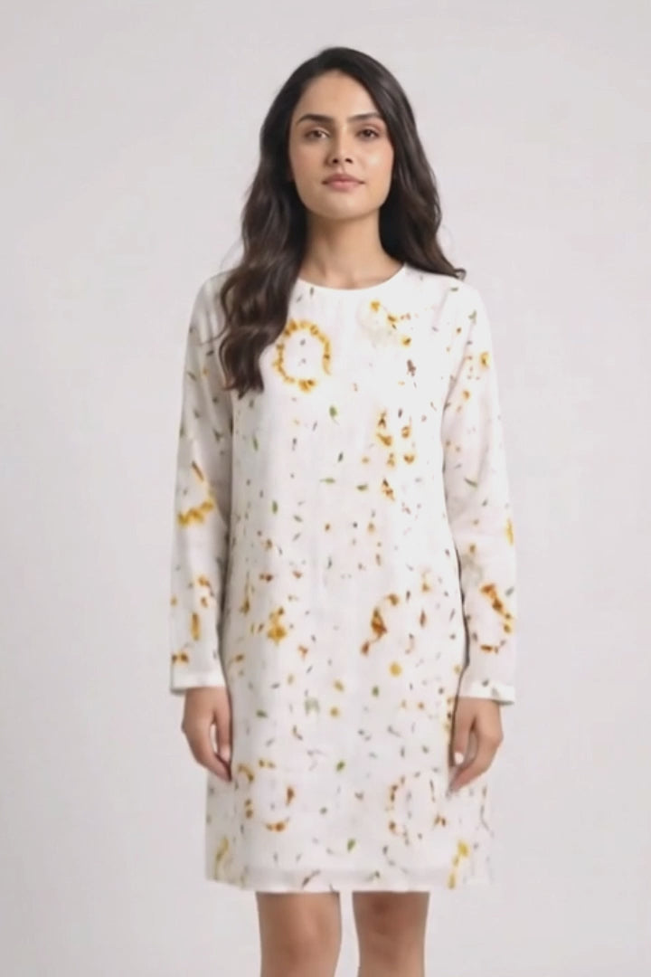 A woman modeling a white long-sleeve shift linen dress with a yellow and orange eco-print, paired with white sneakers, against a light gray studio background.