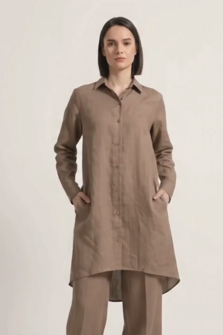 A woman stands against a light gray studio background wearing a loose brown long-sleeve button-front linen  shirt with wide-leg white pants and beige shoes.