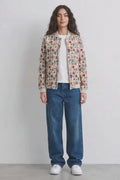 A model stands against a plain light gray wall wearing a cream-colored bomber linen jacket with a dense multicolor floral and botanical print, a white t-shirt, loose-fit blue jeans, and white sneakers.