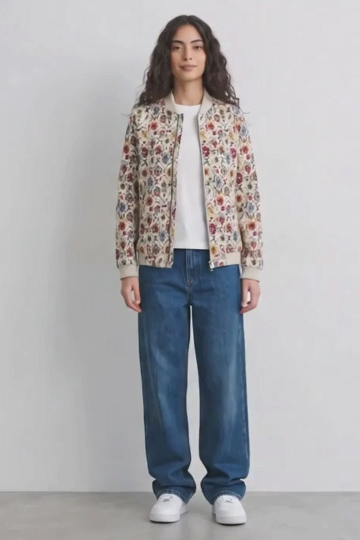 A model stands against a plain light gray wall wearing a cream-colored bomber linen jacket with a dense multicolor floral and botanical print, a white t-shirt, loose-fit blue jeans, and white sneakers.