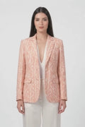 A woman stands against a plain white studio background wearing a peach-pink linen blazer with a white ornamental pattern, white wide-leg pants, and white heeled sandals.