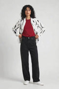 A model stands against a light gray studio background wearing a white hemp bomber jacket with black abstract spots, a red t-shirt, black wide-leg jeans, and white sneakers.