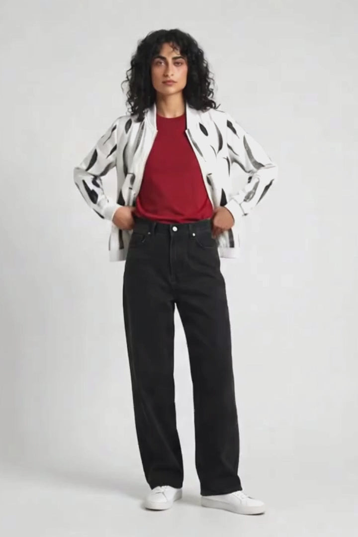 A model stands against a light gray studio background wearing a white hemp bomber jacket with black abstract spots, a red t-shirt, black wide-leg jeans, and white sneakers.