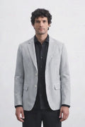 A man modeling a light gray and white vertically striped linen blazer over a black shirt and black trousers, standing against a plain white studio background.