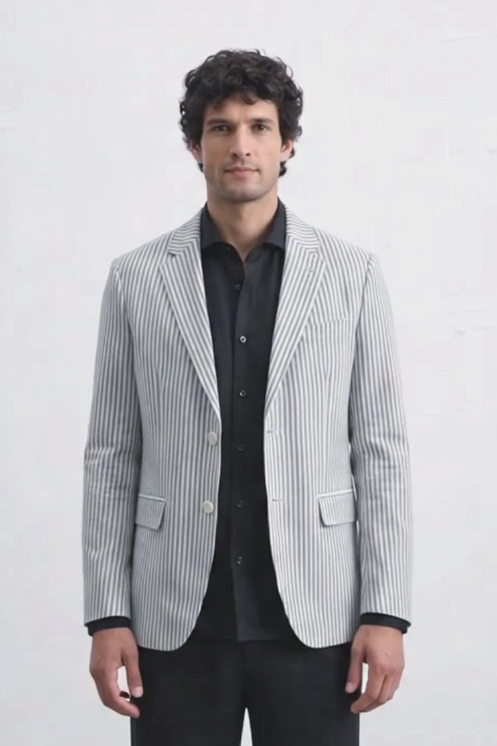 A man modeling a light gray and white vertically striped linen blazer over a black shirt and black trousers, standing against a plain white studio background.