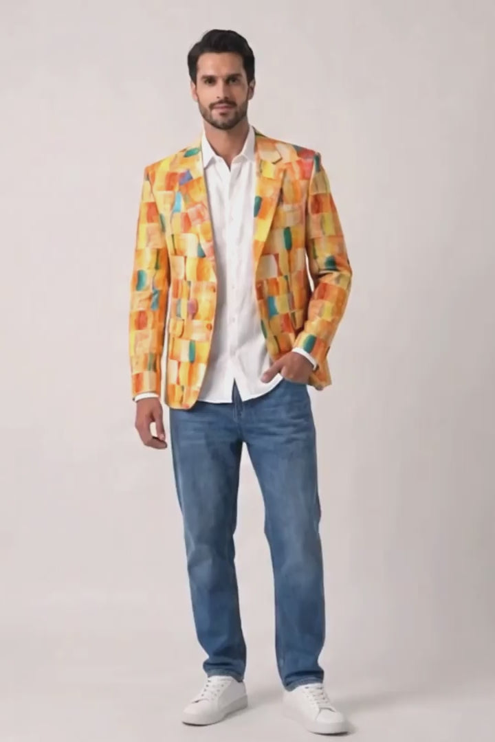 A man modeling a vibrant yellow, orange, and green abstract-patterned hemp blazer over a white button-down shirt, paired with blue jeans and white sneakers, against a plain light background.