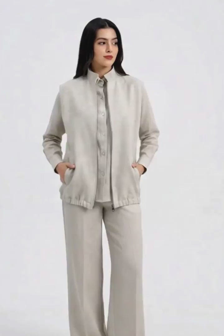 A woman modeling a light beige sleeveless zip-up linen vest with a high stand-up collar and front pockets, set against a plain light gray studio background.