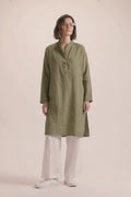 A woman modeling a long-sleeved olive green linen tunic with a split neckline and button placket, paired with white wide-leg trousers and white sneakers, set against a solid pale pink studio background.