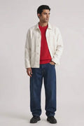 A man modeling a white button-up linen overshirt over a red crewneck sweater, paired with loose blue jeans and black shoes, against a plain light gray studio background.