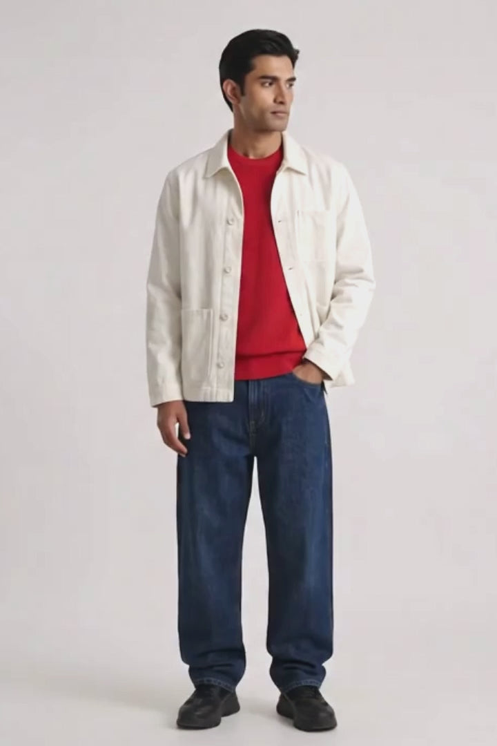 A man modeling a white button-up linen overshirt over a red crewneck sweater, paired with loose blue jeans and black shoes, against a plain light gray studio background.