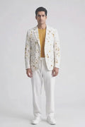 A man modeling a white blazer with a yellow and orange floral pattern, worn over a mustard yellow t-shirt and paired with white trousers and white sneakers, against a plain white studio background.