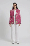 A model stands against a light gray studio background wearing a pink floral silk blazer over a white button-up shirt and white wide-leg trousers, with white sneakers.