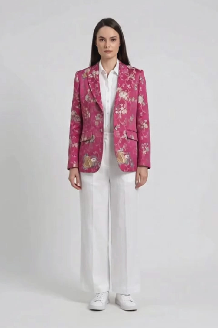 A model stands against a light gray studio background wearing a pink floral silk blazer over a white button-up shirt and white wide-leg trousers, with white sneakers.