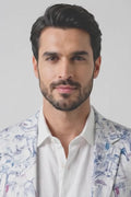 A man modeling a white linen blazer with a blue and gray toile-style print, worn over a white shirt and blue jeans, standing against a light gray studio background.