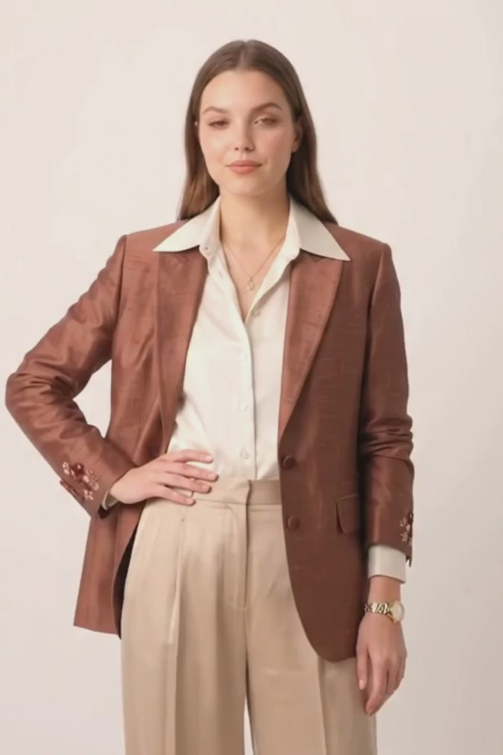 A model stands against a light gray studio background wearing a metallic bronze-brown silk blazer over a cream collared shirt, paired with wide-leg beige trousers and holding a small metallic clutch.