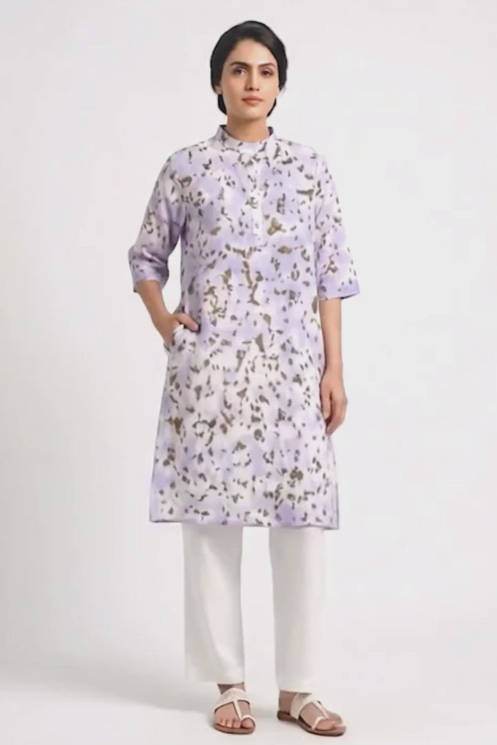 A model wearing a lavender cotton eco-print kurta featuring natural marigold leaf motifs in earthy golden tones. The straight-fit kurta has a mandarin collar, three-quarter sleeves, a short button placket, and side pockets. It is paired with off-white straight pants and white braided sandals against a clean white background.