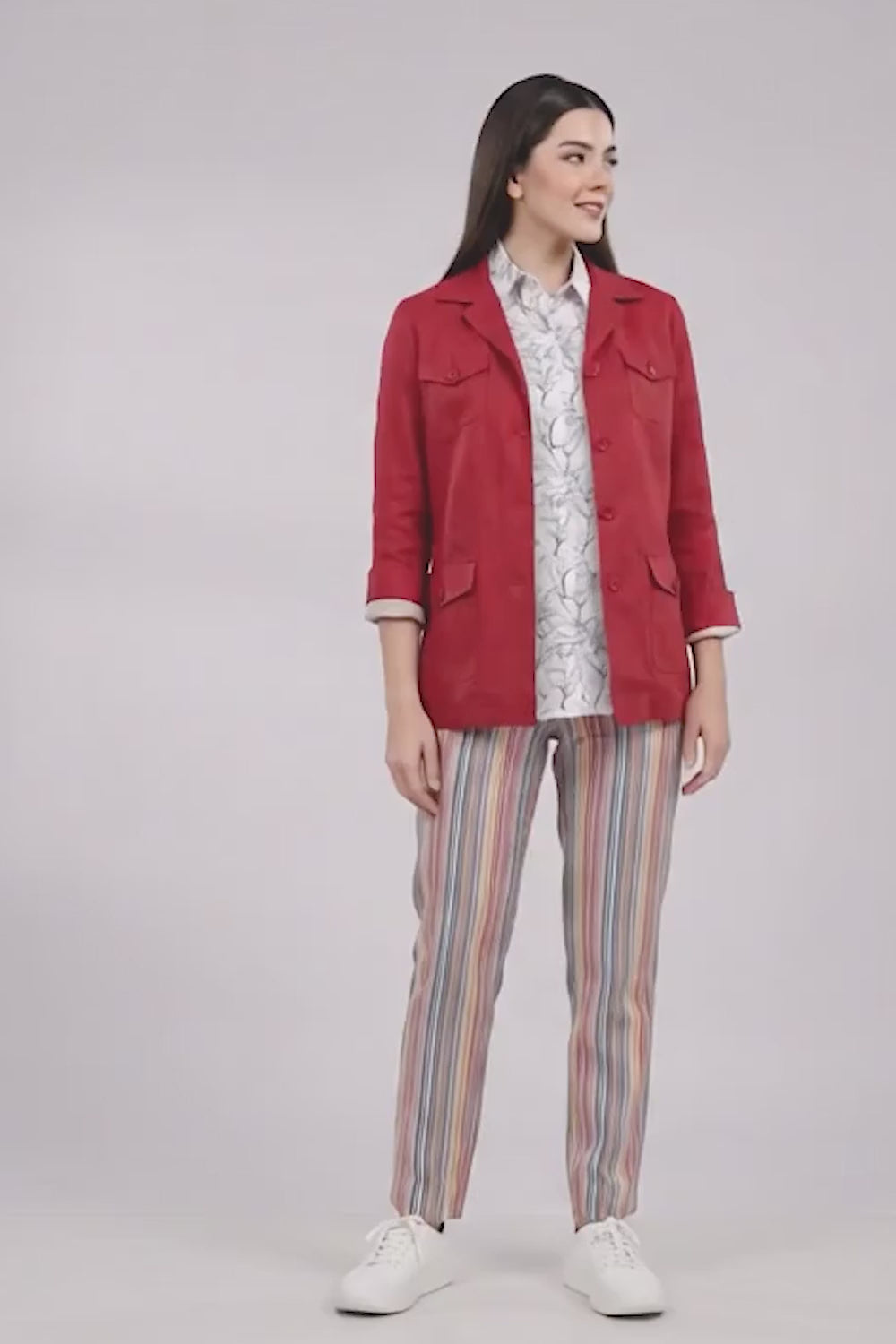 A woman stands against a light gray studio background wearing a red cropped linen jacket over a white patterned shirt, with vertically striped pants in pink, blue, and orange, and white sneakers.