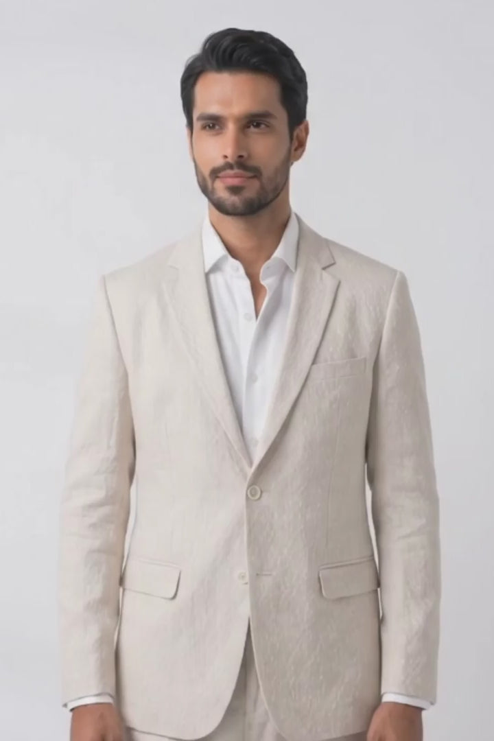 A man modeling a light beige linen suit with a white dress shirt and dark brown loafers, standing against a plain white studio background.