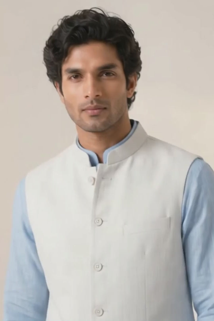 A promotional product video showcasing a man wearing a light grey tailored linen vest over a pale blue kurta and white trousers, featuring medium shots adjusting the vest, full-length walking shots, and close-up details of the fine fabric texture and pocket stitching against a neutral studio backdrop.