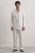 A man modeling a light cream hemp suit consisting of a blazer and trousers, worn over a white shirt and black sandals, standing against a white textured wall.