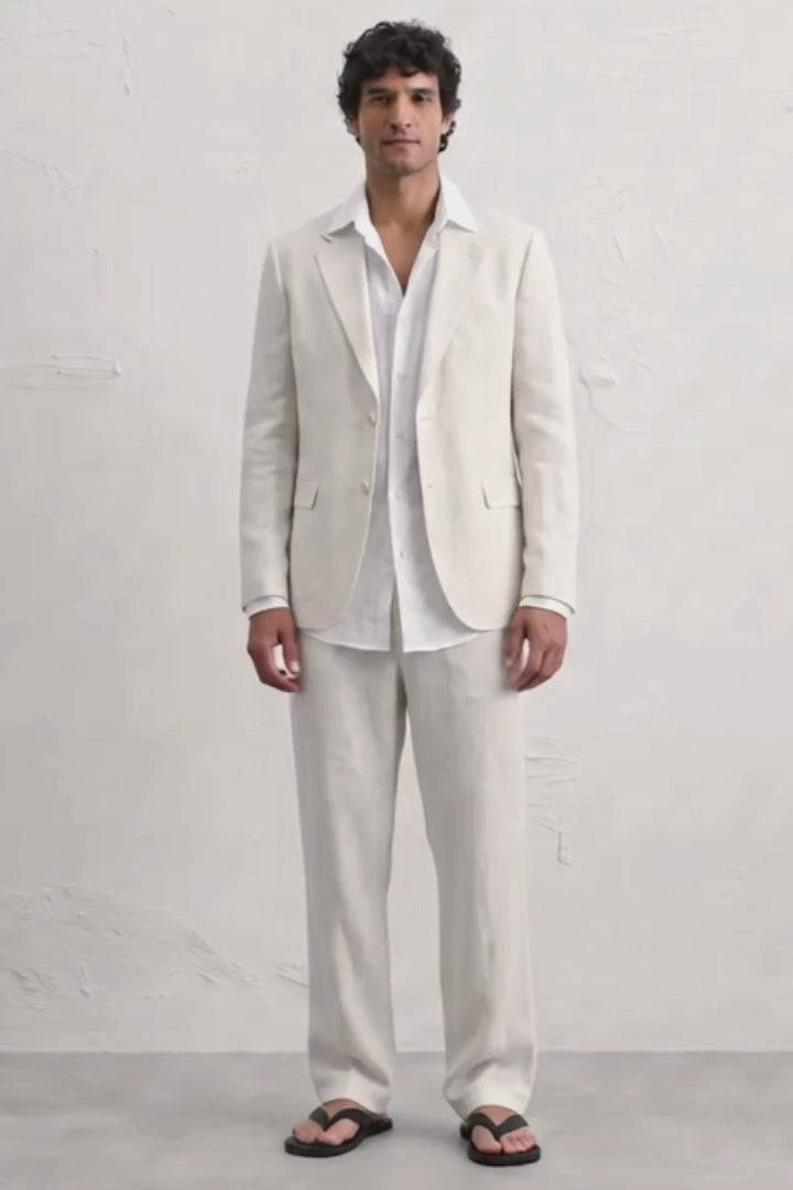 A man modeling a light cream hemp suit consisting of a blazer and trousers, worn over a white shirt and black sandals, standing against a white textured wall.
