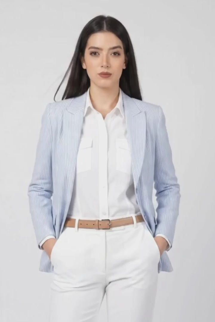 A woman modeling a light blue linen blazer, white shirt, white trousers, and white heels against a light gray studio background.