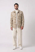 A man modeling a beige and white patterned hemp jacket over a cream turtleneck and cream trousers, standing against a light gray studio background.