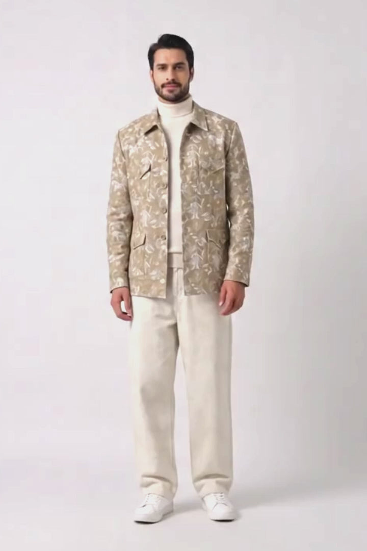 A man modeling a beige and white patterned hemp jacket over a cream turtleneck and cream trousers, standing against a light gray studio background.