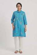 A woman modeling a bright blue kurta with a subtle gold and green floral pattern, paired with white wide-leg trousers and brown sandals, against a plain white studio background.